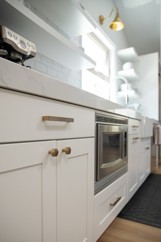 This is an example of a mid-sized transitional galley eat-in kitchen in San Diego with a farmhouse sink, shaker cabinets, white cabinets, quartz benchtops, white splashback, ceramic splashback, stainless steel appliances, light hardwood floors, a peninsula, beige floor and white benchtop.