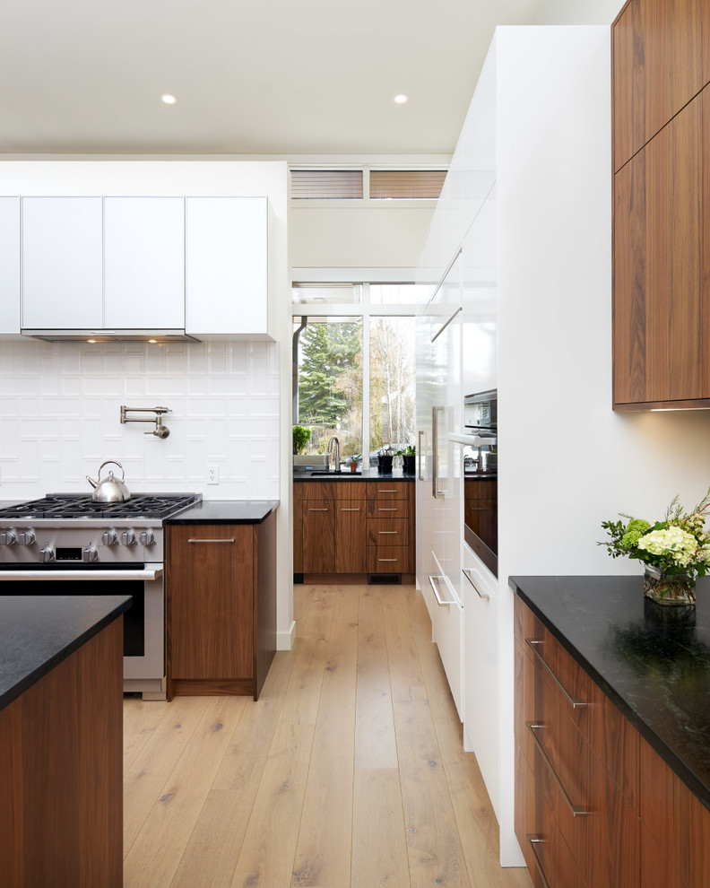 Inspiration for a modern l-shaped light wood floor open concept kitchen remodel in Calgary with flat-panel cabinets, dark wood cabinets, soapstone countertops, white backsplash, stainless steel appliances, an island, black countertops and porcelain backsplash