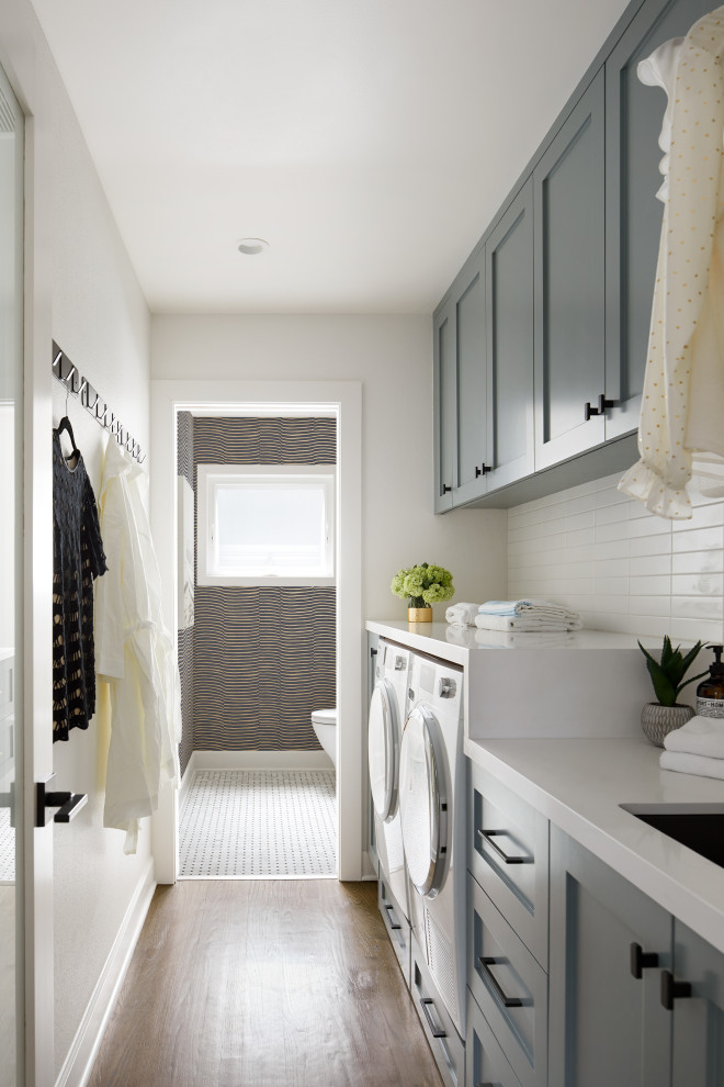 Idéer för en klassisk vita linjär tvättstuga enbart för tvätt, med en undermonterad diskho, skåp i shakerstil, grå skåp, vita väggar, mellanmörkt trägolv, en tvättmaskin och torktumlare bredvid varandra och brunt golv
