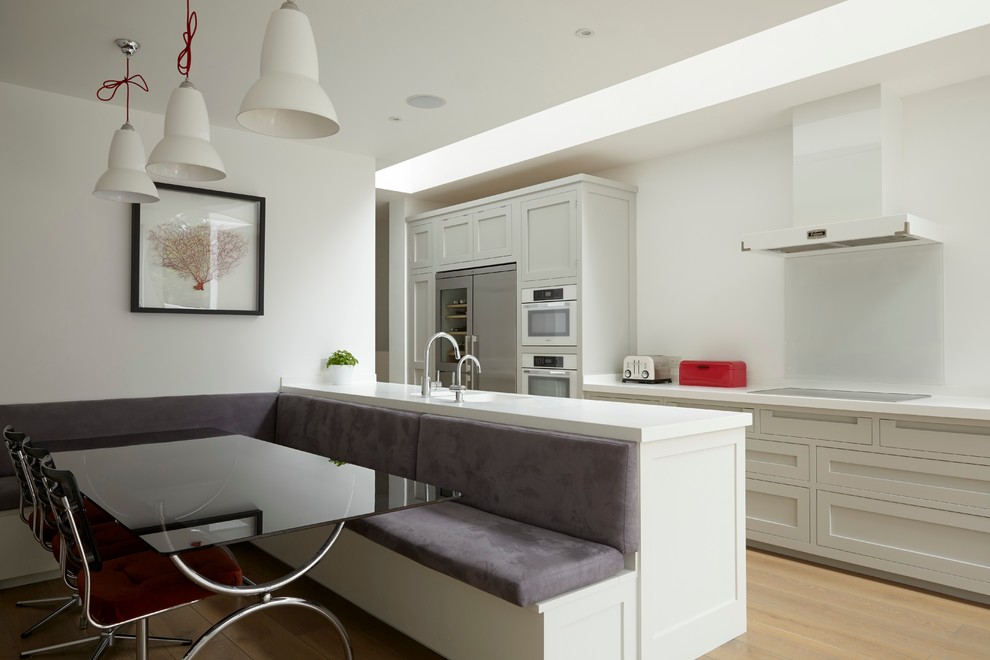 Inspiration for a mid-sized contemporary galley eat-in kitchen in London with a double-bowl sink, shaker cabinets, white cabinets, solid surface benchtops, white splashback, glass sheet splashback, panelled appliances, light hardwood floors and with island.