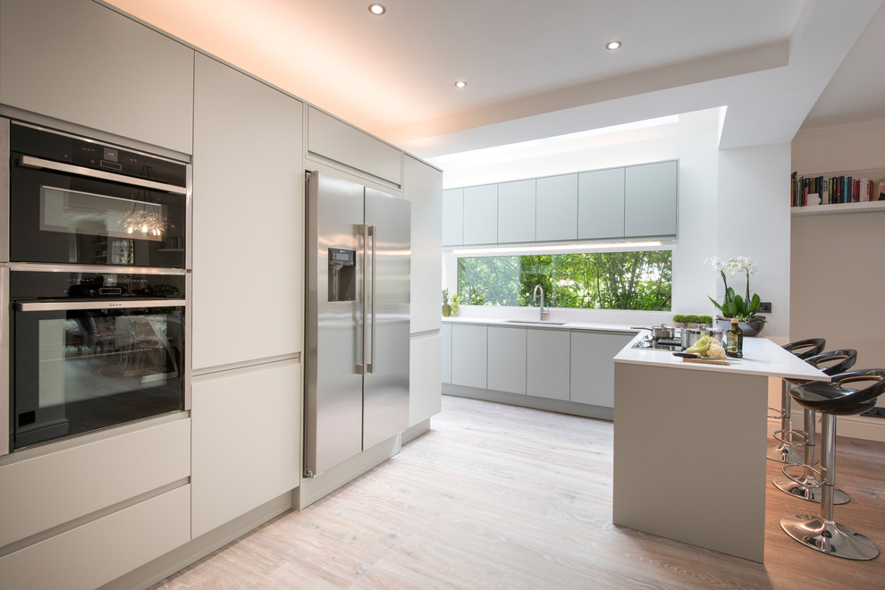 Design ideas for a contemporary u-shaped kitchen in London with an undermount sink, flat-panel cabinets, grey cabinets, stainless steel appliances, light hardwood floors, a peninsula, beige floor and white benchtop.
