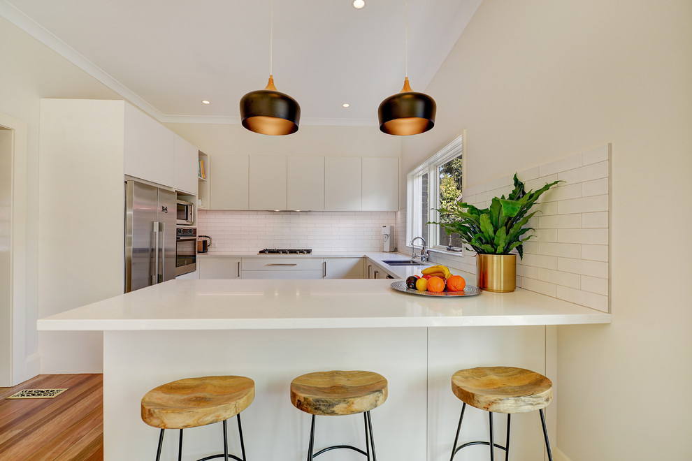 Mid-sized modern u-shaped eat-in kitchen in Sydney with an undermount sink, flat-panel cabinets, white cabinets, quartz benchtops, white splashback, subway tile splashback, stainless steel appliances, light hardwood floors, a peninsula, brown floor and white benchtop.