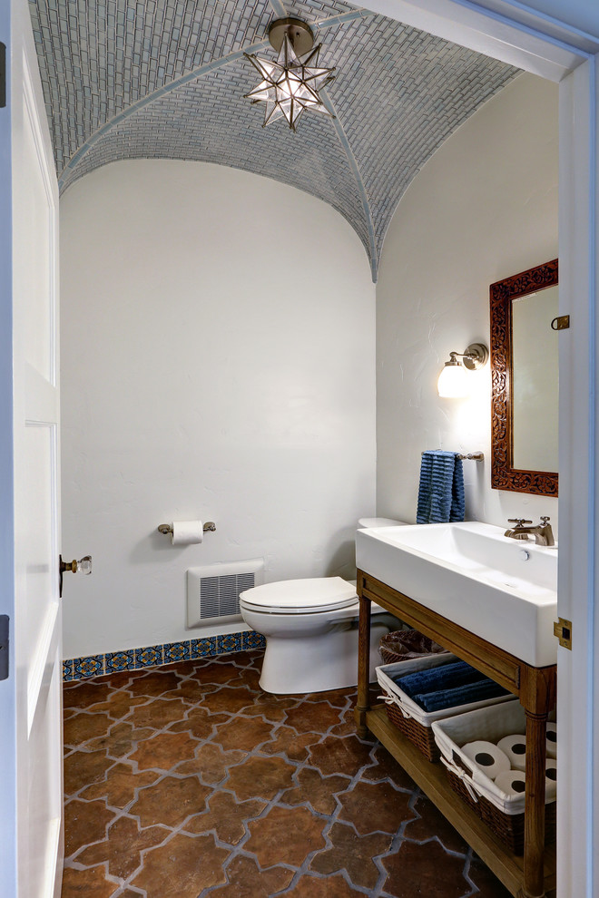 Mid-sized mediterranean powder room in Los Angeles with open cabinets, a trough sink, medium wood cabinets, a two-piece toilet, white walls, terra-cotta floors, solid surface benchtops and multi-coloured floor.