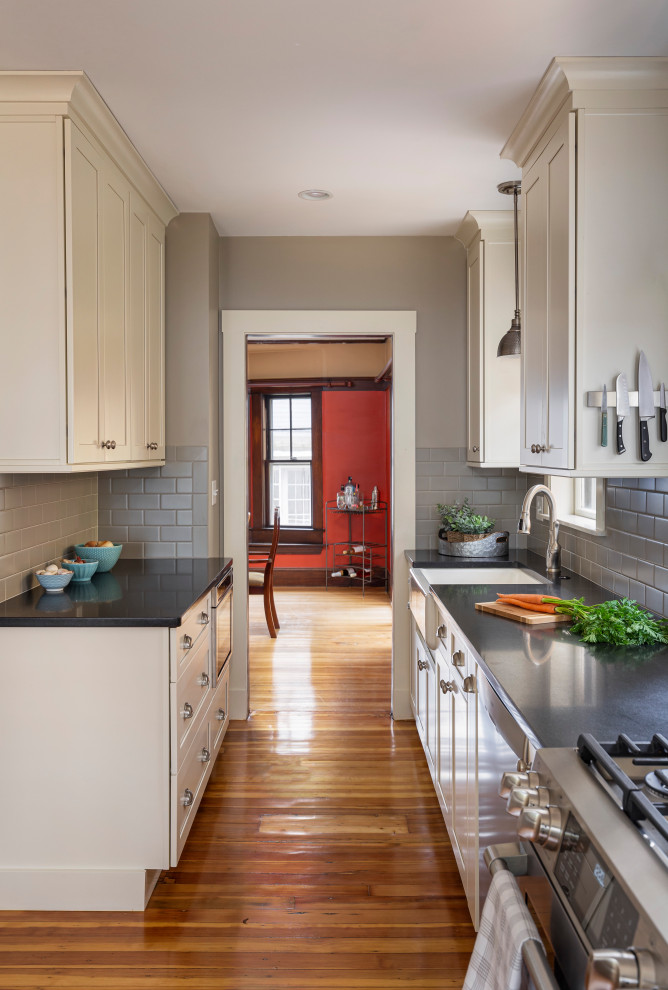 This is an example of a mid-sized transitional galley kitchen pantry in Boston with a farmhouse sink, shaker cabinets, white cabinets, granite benchtops, grey splashback, subway tile splashback, stainless steel appliances, light hardwood floors, no island and black benchtop.