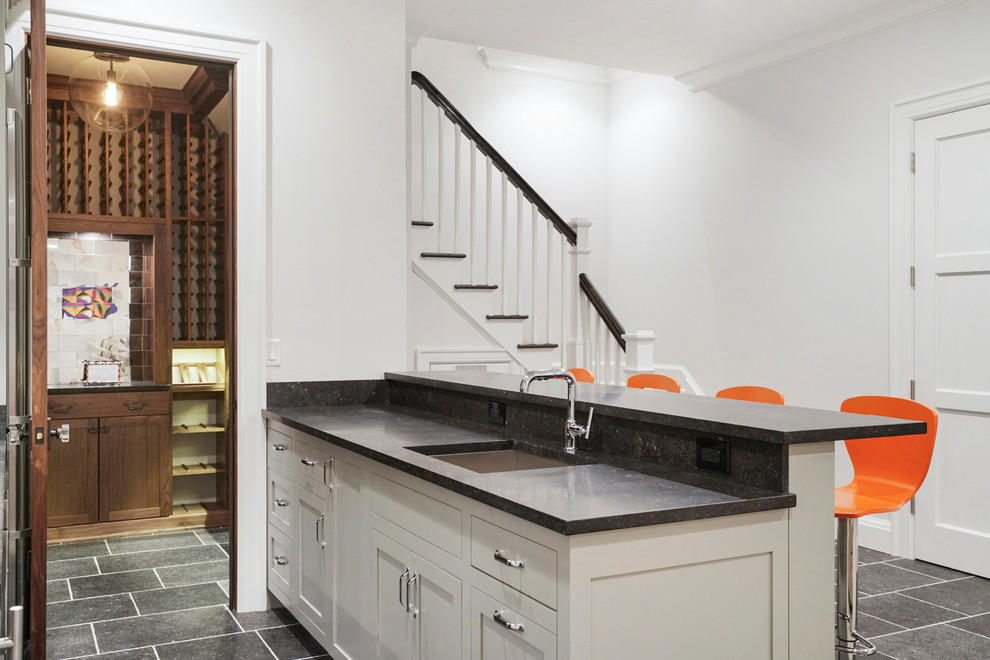 Inspiration for an expansive transitional galley seated home bar in New York with an undermount sink, shaker cabinets, grey cabinets, limestone benchtops, multi-coloured splashback and limestone floors.