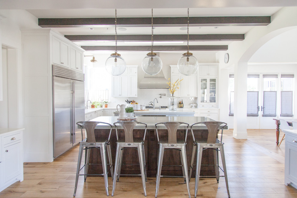 Photo of a large transitional l-shaped eat-in kitchen in Phoenix with a farmhouse sink, white cabinets, white splashback, stainless steel appliances, light hardwood floors, with island, stainless steel benchtops, shaker cabinets and porcelain splashback.