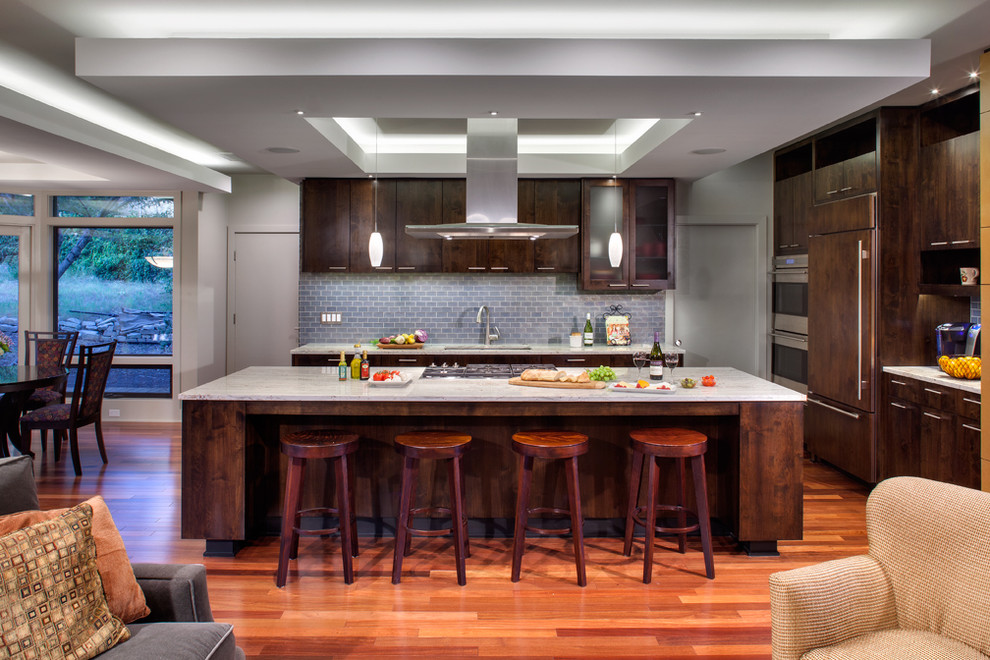 Contemporary l-shaped open plan kitchen in Austin with dark wood cabinets, grey splashback, panelled appliances, flat-panel cabinets, granite benchtops, glass tile splashback, medium hardwood floors, with island and an undermount sink.