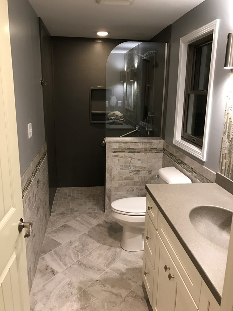 Example of a mid-sized transitional master gray tile and porcelain tile porcelain tile walk-in shower design with recessed-panel cabinets, white cabinets, a two-piece toilet, gray walls, an integrated sink and solid surface countertops