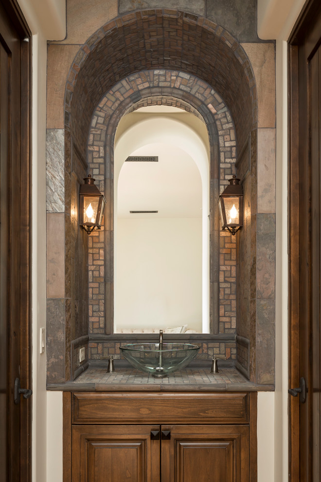 This is an example of a mid-sized mediterranean powder room in Phoenix with raised-panel cabinets, dark wood cabinets, multi-coloured tile, mosaic tile, white walls, tile benchtops and a vessel sink.