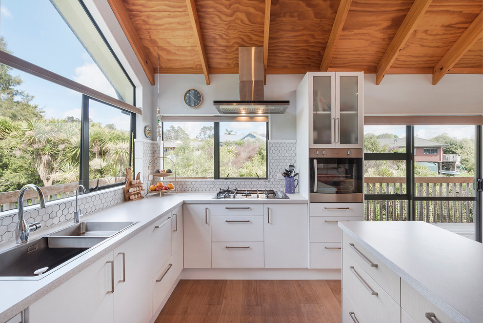 This is an example of a large contemporary u-shaped open plan kitchen in Auckland with flat-panel cabinets, white cabinets, a double-bowl sink, quartz benchtops, multi-coloured splashback, mosaic tile splashback, white appliances, with island and brown floor.
