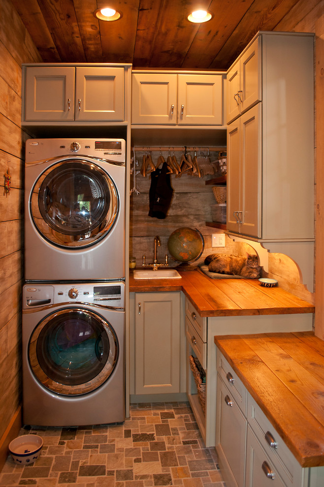 This is an example of a mid-sized country galley utility room in Other with a drop-in sink, shaker cabinets, grey cabinets, wood benchtops, brown walls, ceramic floors and a stacked washer and dryer.