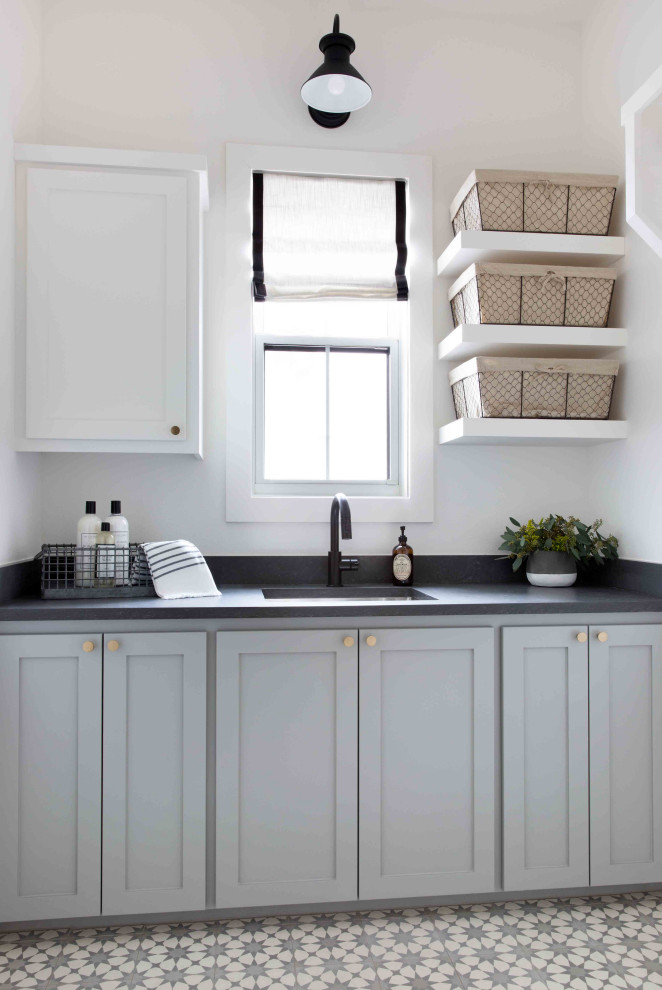 Inspiration for a mid-sized country single-wall dedicated laundry room in Austin with an undermount sink, shaker cabinets, grey cabinets, white walls, multi-coloured floor and black benchtop.