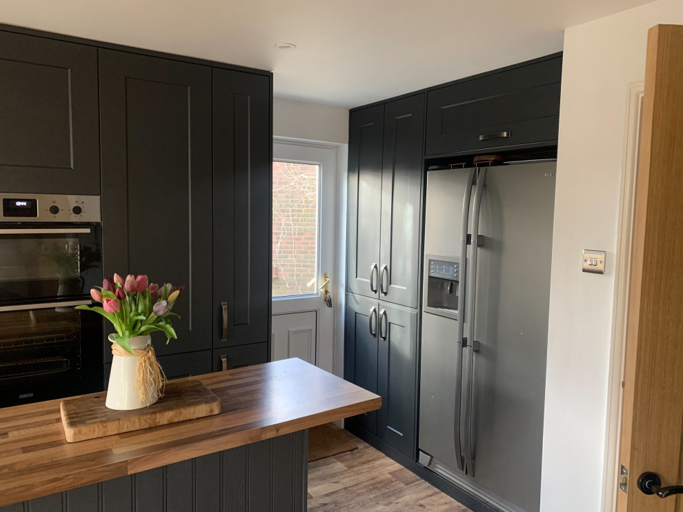 Design ideas for a medium sized classic u-shaped enclosed kitchen in Surrey with a double-bowl sink, recessed-panel cabinets, black cabinets, laminate countertops, black appliances, medium hardwood flooring, an island, brown floors and brown worktops.