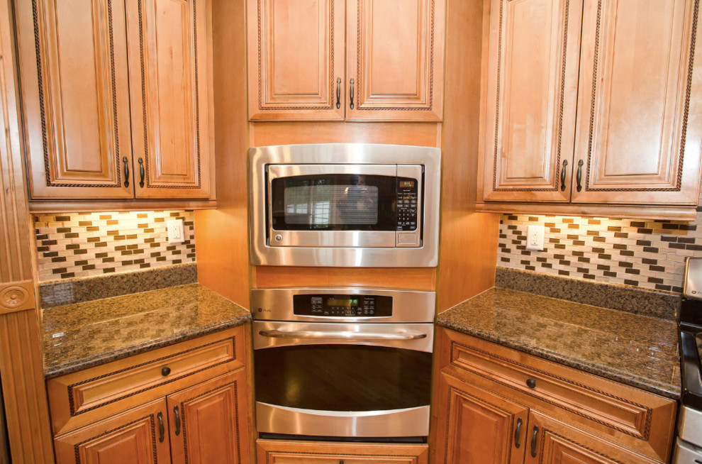 Example of a medium tone wood floor and brown floor kitchen design with a double-bowl sink, glass-front cabinets, medium tone wood cabinets, granite countertops, black backsplash, ceramic backsplash, stainless steel appliances, an island and brown countertops