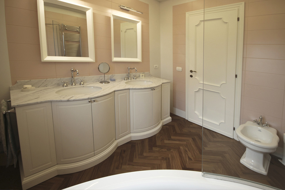 Photo of a large traditional master bathroom in Other with furniture-like cabinets, beige cabinets, a corner tub, a two-piece toilet, pink tile, ceramic tile, pink walls, dark hardwood floors, a drop-in sink, marble benchtops, brown floor and white benchtops.