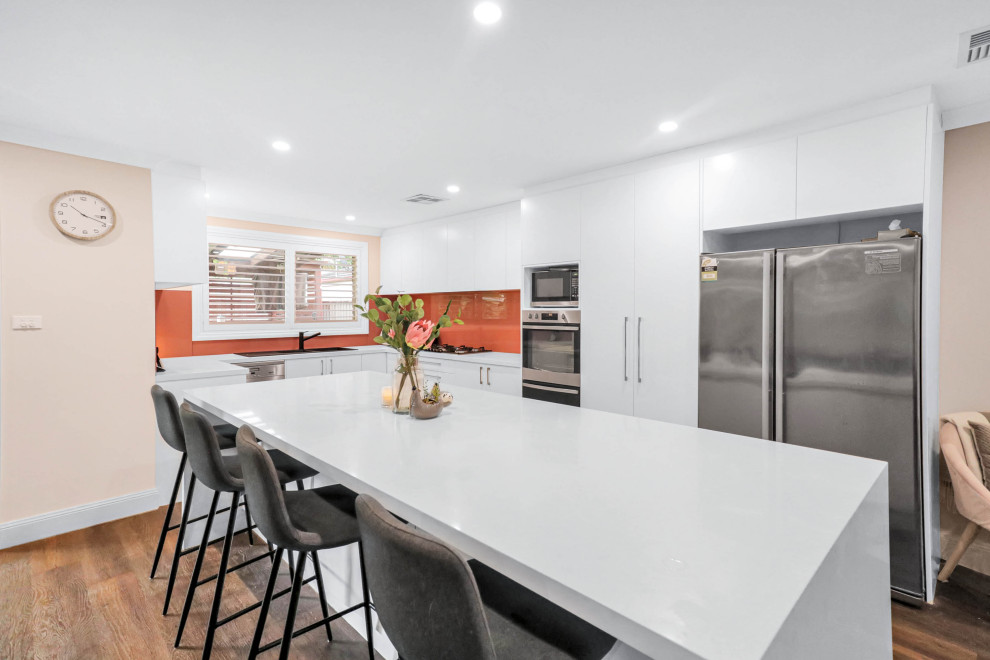 Inspiration for a large modern galley open plan kitchen in Sydney with a double-bowl sink, flat-panel cabinets, white cabinets, quartz benchtops, orange splashback, glass sheet splashback, stainless steel appliances, vinyl floors, with island, brown floor and white benchtop.