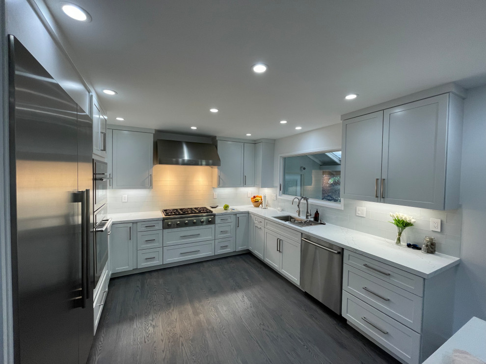 Bild på ett stort funkis vit vitt kök, med en dubbel diskho, skåp i shakerstil, vita skåp, bänkskiva i kvarts, stänkskydd i glaskakel, rostfria vitvaror, mörkt trägolv, en köksö och grått golv