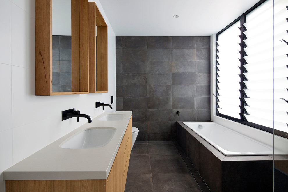 This is an example of a large contemporary kids bathroom in Sydney with flat-panel cabinets, medium wood cabinets, a drop-in tub, an open shower, a one-piece toilet, gray tile, ceramic tile, ceramic floors, an undermount sink, engineered quartz benchtops, grey floor and white benchtops.