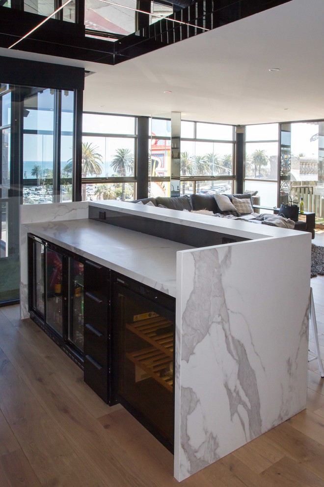 This is an example of a large modern galley seated home bar in Melbourne with flat-panel cabinets, a drop-in sink, black cabinets, tile benchtops, grey splashback, mosaic tile splashback, medium hardwood floors, brown floor and white benchtop.