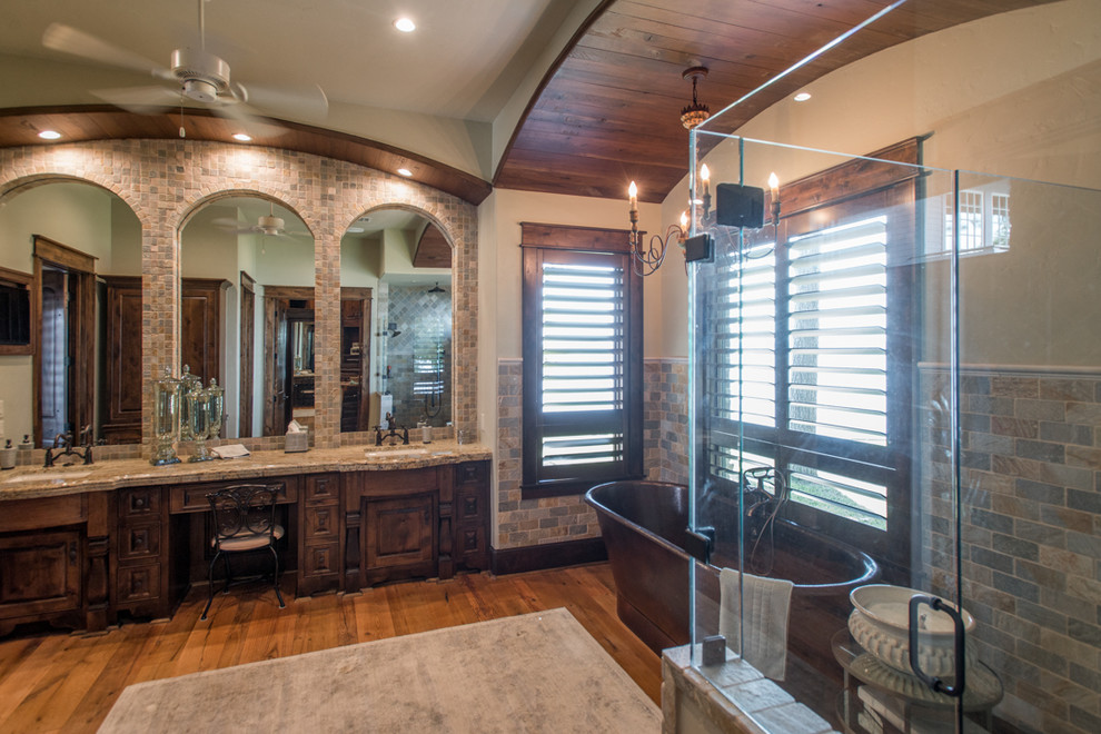 Inspiration for a large country master bathroom in Houston with recessed-panel cabinets, distressed cabinets, a freestanding tub, a corner shower, beige tile, stone tile, beige walls, medium hardwood floors, an undermount sink, granite benchtops, brown floor and a hinged shower door.
