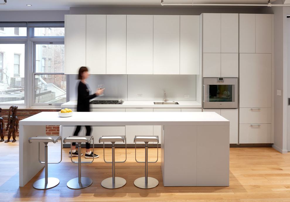 This is an example of an industrial kitchen in DC Metro with a single-bowl sink, flat-panel cabinets, white cabinets, white splashback, glass sheet splashback, panelled appliances, light hardwood floors, with island and white benchtop.