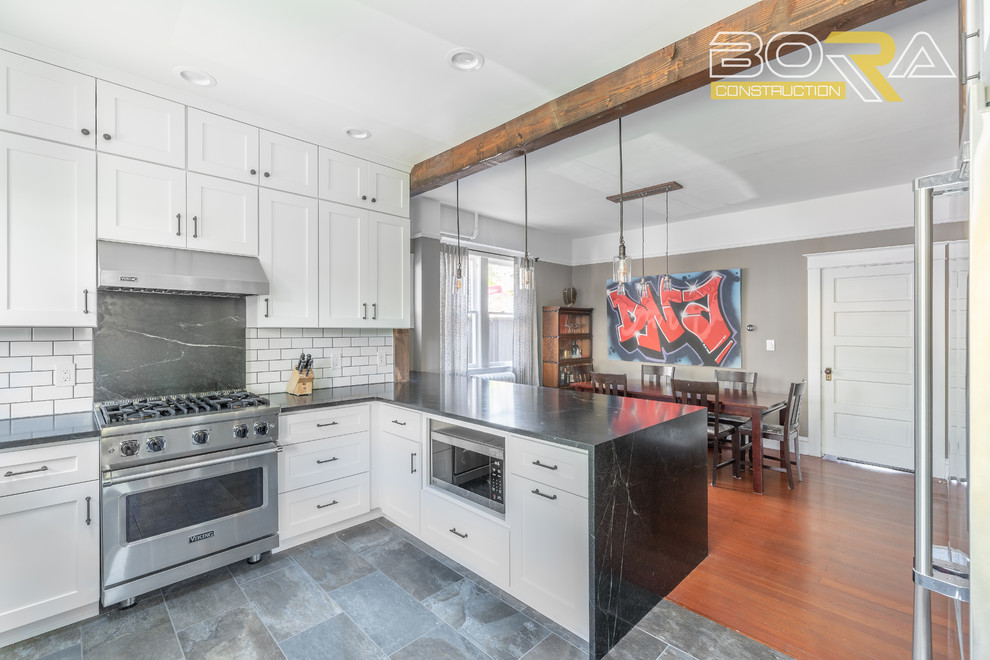 Example of a mid-sized transitional u-shaped ceramic tile and gray floor eat-in kitchen design in Seattle with a farmhouse sink, shaker cabinets, white cabinets, quartz countertops, white backsplash, ceramic backsplash, stainless steel appliances, a peninsula and black countertops