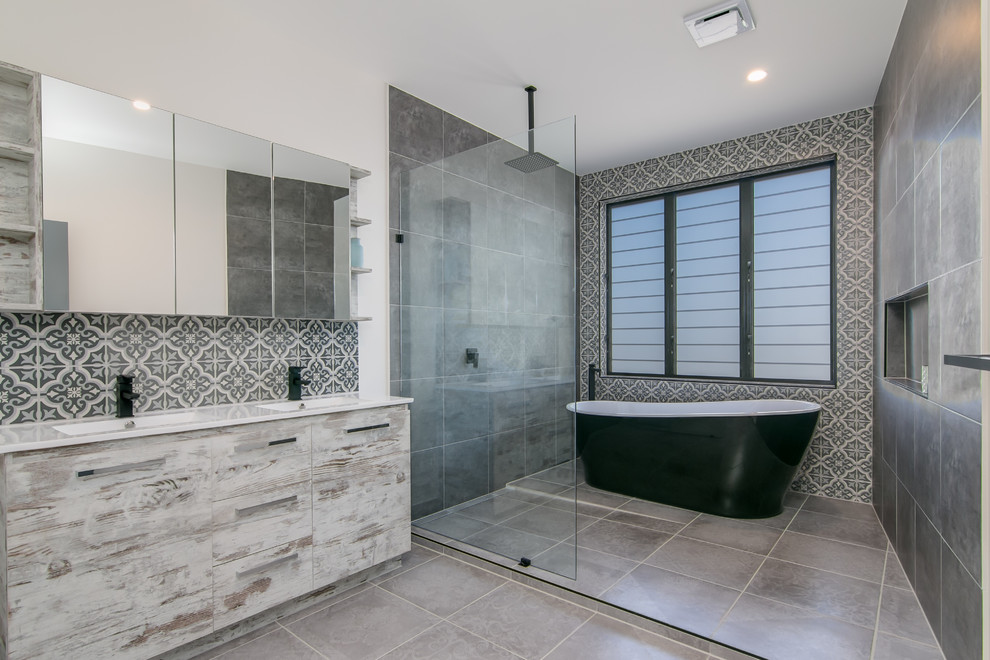 This is an example of a large contemporary master bathroom in Cairns with flat-panel cabinets, distressed cabinets, a freestanding tub, an open shower, a one-piece toilet, gray tile, ceramic tile, white walls, ceramic floors, an integrated sink and solid surface benchtops.