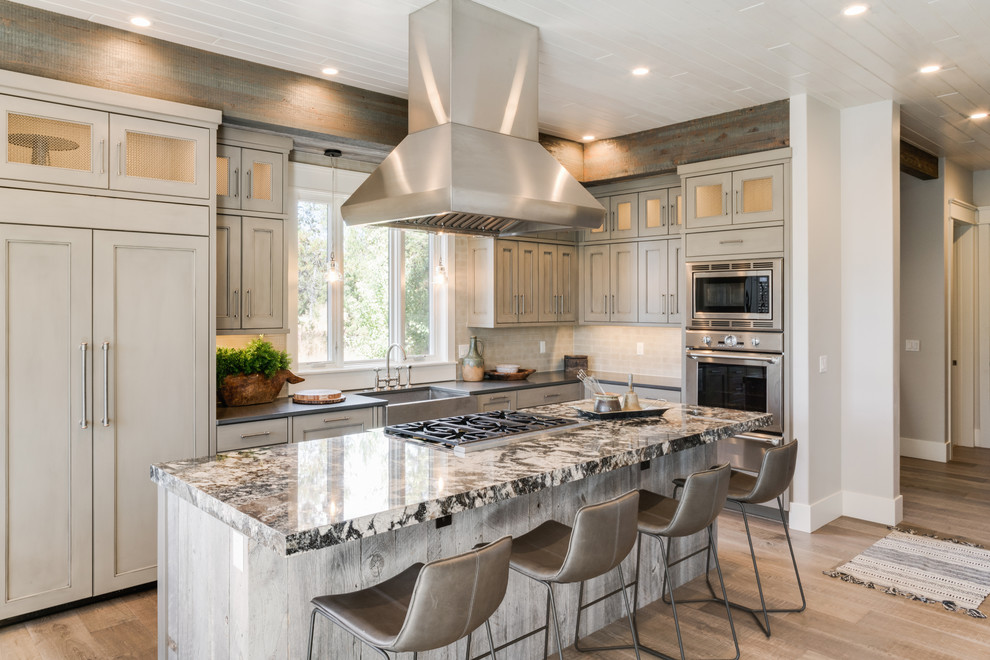Photo of a country l-shaped kitchen in Boise with a farmhouse sink, recessed-panel cabinets, beige cabinets, beige splashback, stainless steel appliances, light hardwood floors, with island, multi-coloured benchtop and granite benchtops.
