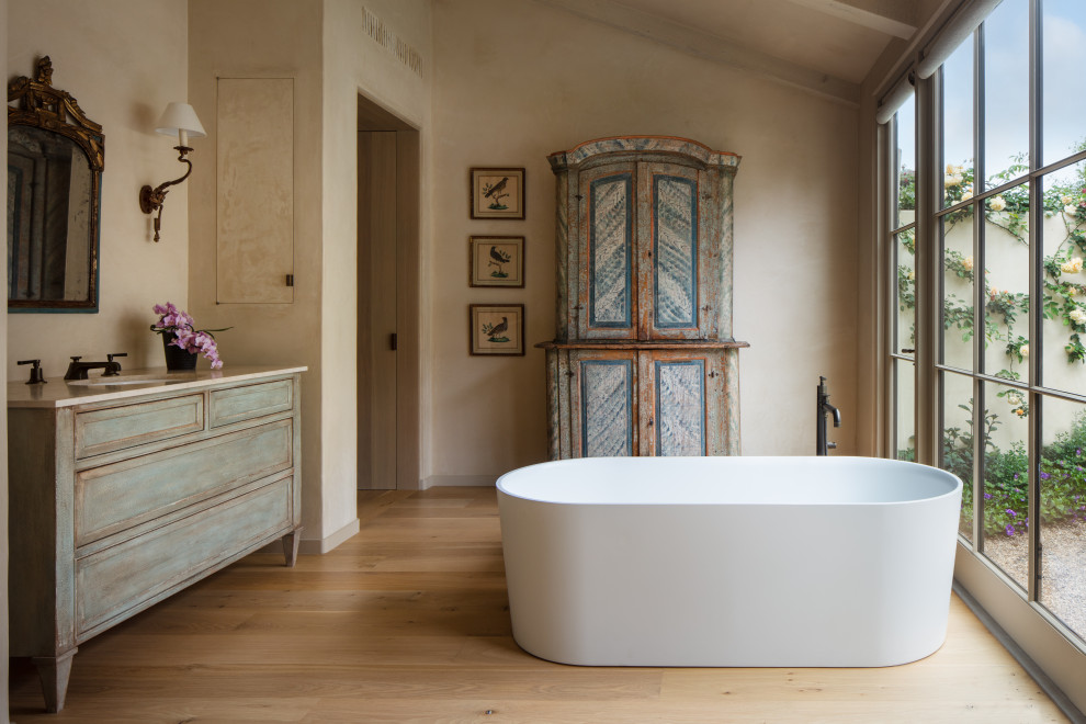 Foto på ett stort medelhavsstil beige en-suite badrum, med ett fristående badkar, beige väggar, mellanmörkt trägolv, brunt golv, luckor med upphöjd panel, grå skåp och ett undermonterad handfat
