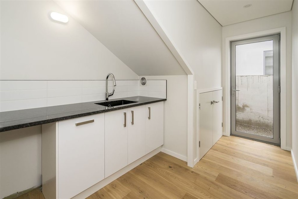 Inspiration for a mid-sized modern single-wall dedicated laundry room in Christchurch with an undermount sink, white cabinets, granite benchtops, white walls and light hardwood floors.