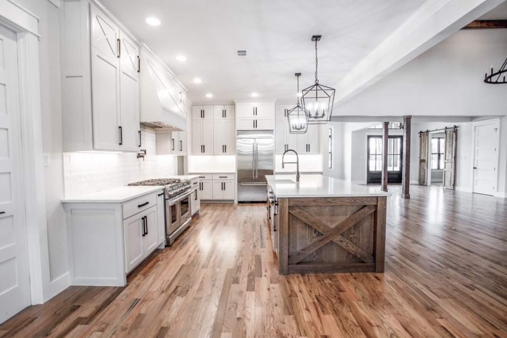 Design ideas for a large country l-shaped open plan kitchen in Dallas with a farmhouse sink, shaker cabinets, white cabinets, quartzite benchtops, white splashback, ceramic splashback, stainless steel appliances, medium hardwood floors, with island, brown floor and white benchtop.
