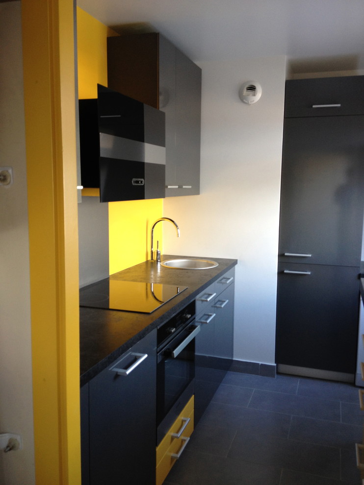 Small modern galley separate kitchen in Lyon with an undermount sink, flat-panel cabinets, yellow cabinets, stainless steel benchtops, yellow splashback, glass sheet splashback, black appliances, ceramic floors, no island, grey floor and black benchtop.