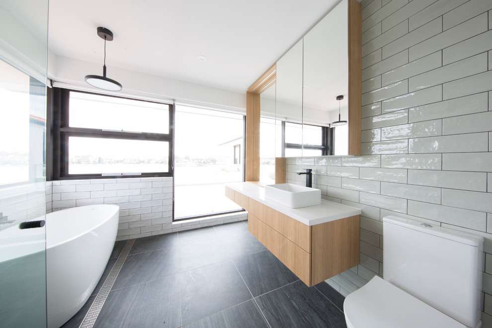 Inspiration for a contemporary 3/4 bathroom in Sydney with flat-panel cabinets, medium wood cabinets, a freestanding tub, a one-piece toilet, white tile, ceramic tile, white walls, a vessel sink, black floor and white benchtops.