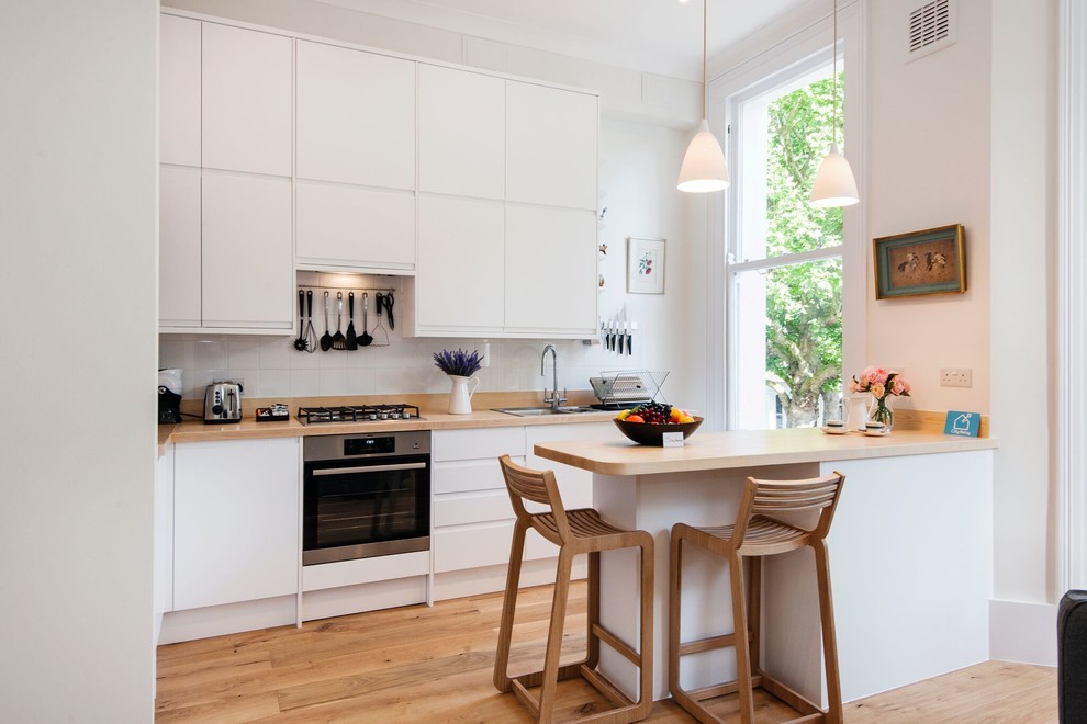 Design ideas for a small contemporary l-shaped kitchen in London with a double-bowl sink, flat-panel cabinets, white cabinets, wood benchtops, white splashback, stainless steel appliances, light hardwood floors, a peninsula, beige floor and beige benchtop.