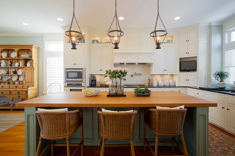This is an example of a large beach style l-shaped eat-in kitchen in Charleston with a drop-in sink, louvered cabinets, white cabinets, wood benchtops, white splashback, ceramic splashback, stainless steel appliances, medium hardwood floors, with island and black benchtop.
