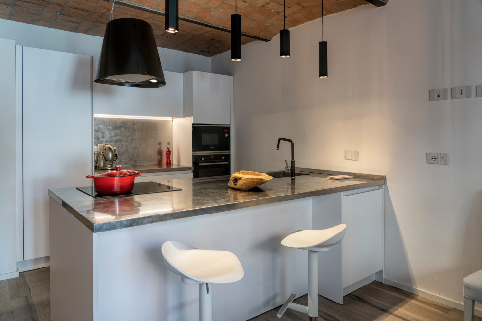 Design ideas for a contemporary galley kitchen in Bologna with an integrated sink, flat-panel cabinets, white cabinets, stainless steel benchtops, metallic splashback, black appliances, medium hardwood floors, a peninsula, brown floor, grey benchtop and vaulted.