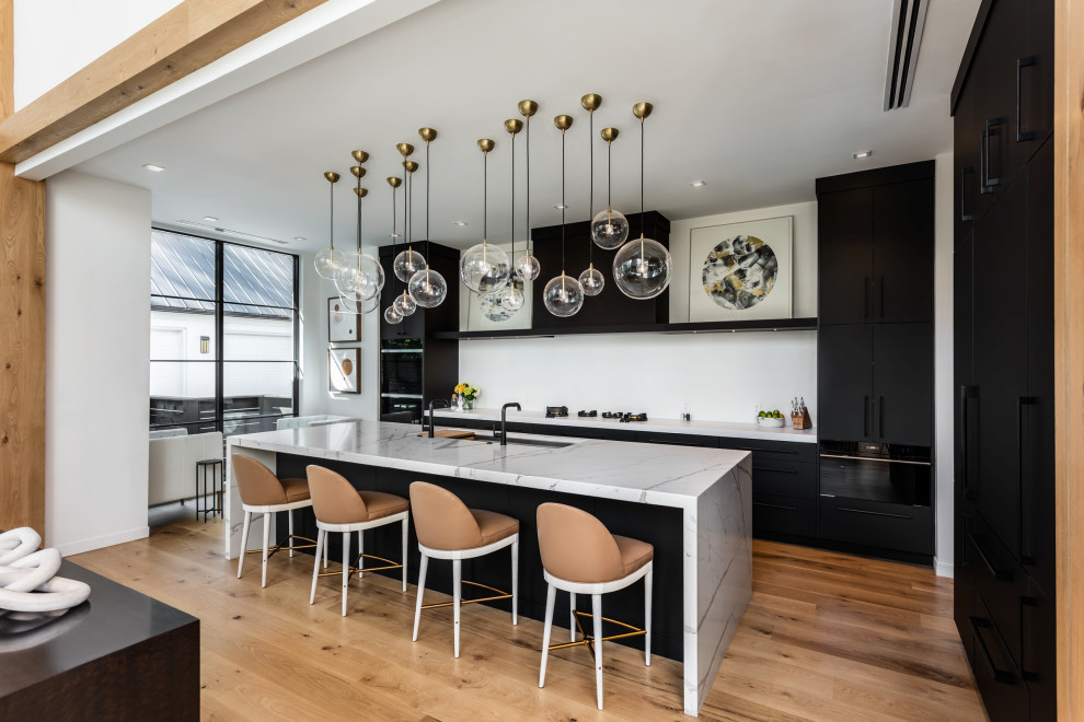 Large contemporary l-shaped open plan kitchen in Other with a submerged sink, flat-panel cabinets, black cabinets, white splashback, integrated appliances, light hardwood flooring, an island, brown floors and white worktops.