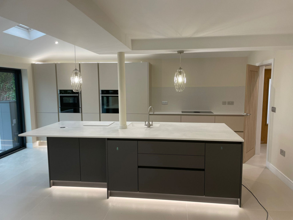 Design ideas for a mid-sized contemporary single-wall open plan kitchen in Other with a drop-in sink, flat-panel cabinets, beige cabinets, marble benchtops, white splashback, marble splashback, black appliances, porcelain floors, with island, beige floor, white benchtop and vaulted.