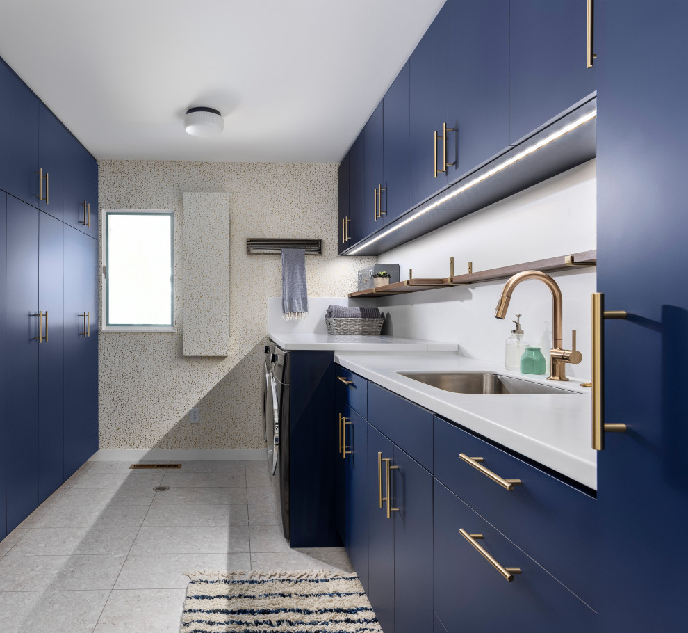 Inspiration for a retro galley separated utility room in Detroit with blue cabinets, a side by side washer and dryer, white worktops, a submerged sink, flat-panel cabinets, engineered stone countertops, white walls, white floors and wallpapered walls.