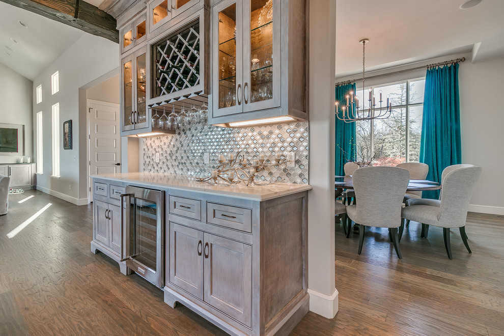 Photo of an expansive country single-wall wet bar in Oklahoma City with no sink, shaker cabinets, light wood cabinets, quartz benchtops, grey splashback, mosaic tile splashback, light hardwood floors and brown floor.