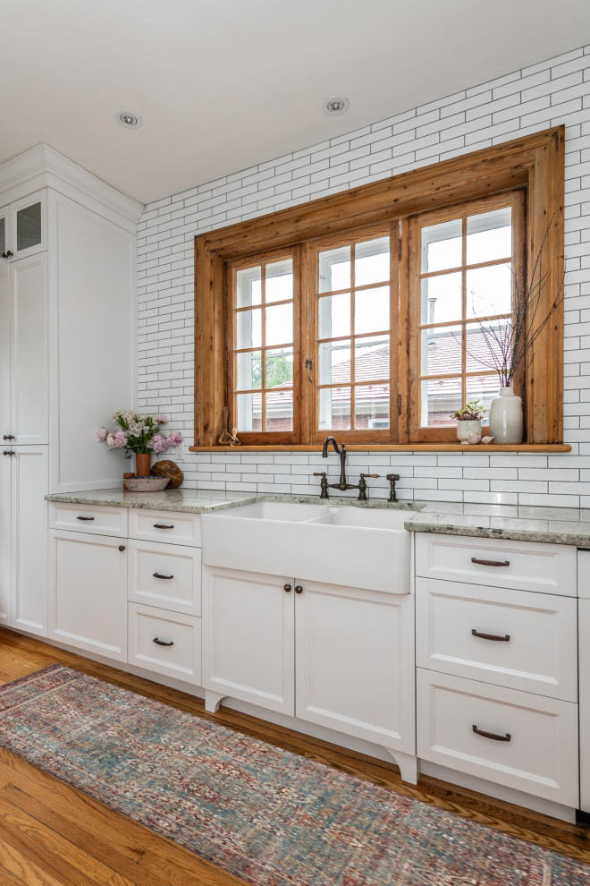 Small transitional galley separate kitchen in Ottawa with a farmhouse sink, shaker cabinets, white cabinets, granite benchtops, white splashback, subway tile splashback, stainless steel appliances, light hardwood floors, with island, brown floor and grey benchtop.
