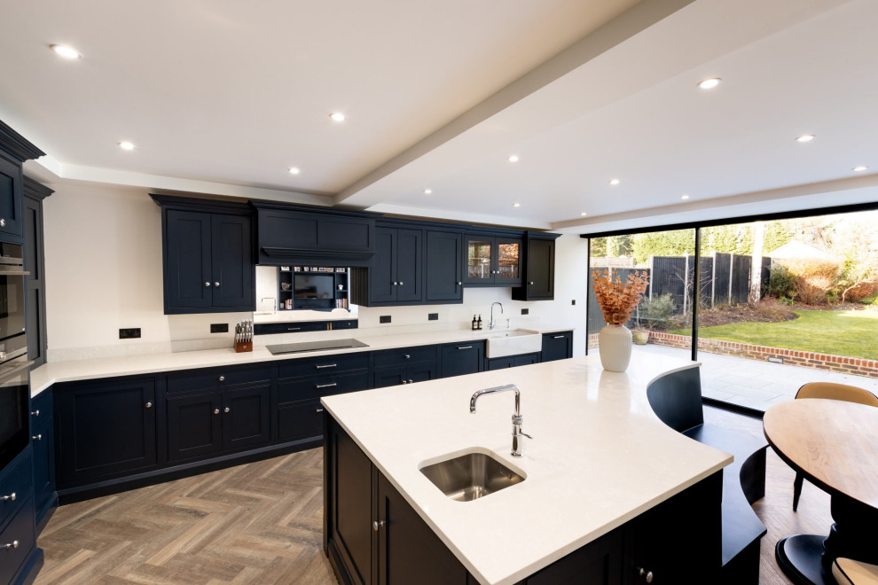 Expansive contemporary l-shaped open plan kitchen in Hertfordshire with a farmhouse sink, shaker cabinets, black cabinets, metallic splashback, glass sheet splashback, black appliances, medium hardwood floors, with island, brown floor and white benchtop.