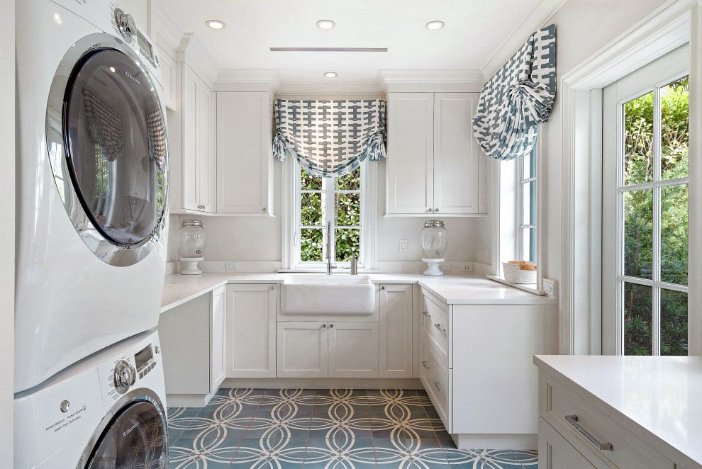 Design ideas for an expansive beach style u-shaped utility room in Miami with a farmhouse sink, recessed-panel cabinets, white cabinets, white walls, a stacked washer and dryer, blue floor and white benchtop.