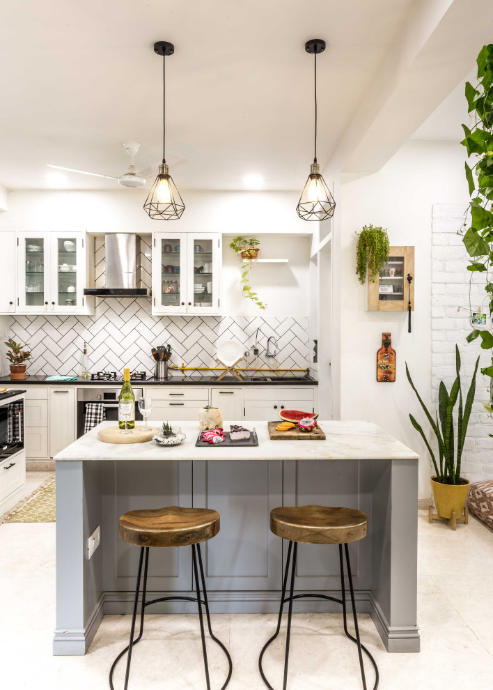 This is an example of an eclectic l-shaped kitchen in Ahmedabad with glass-front cabinets, white cabinets, white splashback, stainless steel appliances, with island, beige floor and black benchtop.