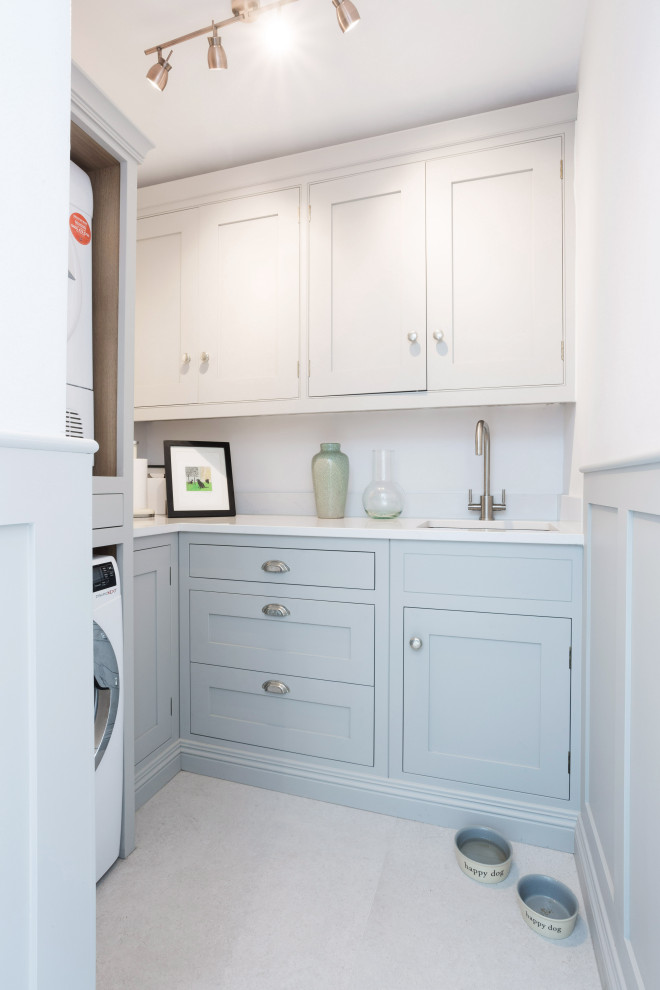 Inspiration for a small country l-shaped utility room in Hertfordshire with an integrated sink, flat-panel cabinets, blue cabinets, quartzite benchtops, white walls, ceramic floors, a stacked washer and dryer, white floor and white benchtop.