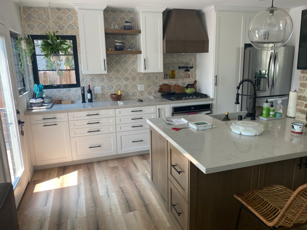 This is an example of a small transitional single-wall open plan kitchen in Los Angeles with a farmhouse sink, shaker cabinets, white cabinets, granite benchtops, brown splashback, ceramic splashback, stainless steel appliances, linoleum floors, with island, multi-coloured floor and beige benchtop.