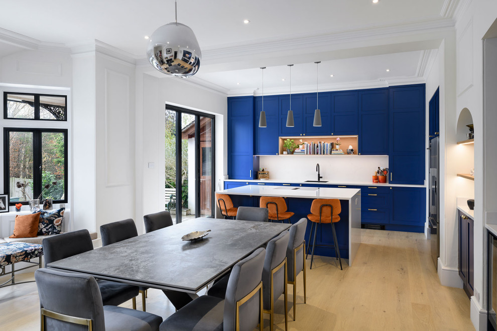 Inspiration for a large transitional l-shaped open plan kitchen in Surrey with medium hardwood floors, a drop-in sink, shaker cabinets, blue cabinets, quartzite benchtops, white splashback, engineered quartz splashback, black appliances, with island and white benchtop.