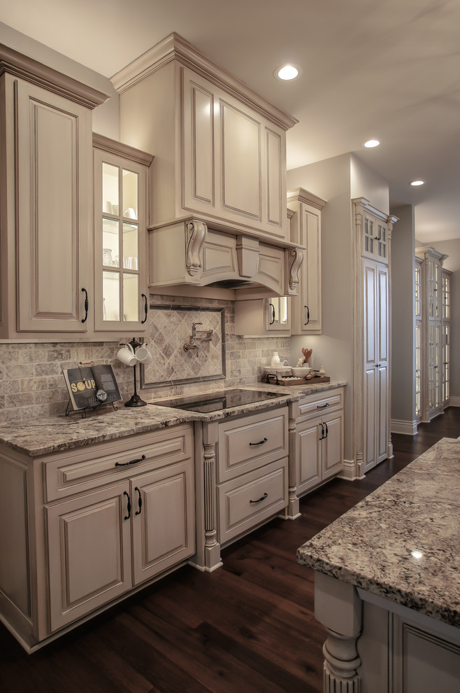 Large traditional l-shaped eat-in kitchen in Omaha with a farmhouse sink, raised-panel cabinets, distressed cabinets, granite benchtops, multi-coloured splashback, stone tile splashback, panelled appliances, dark hardwood floors, with island, brown floor and beige benchtop.