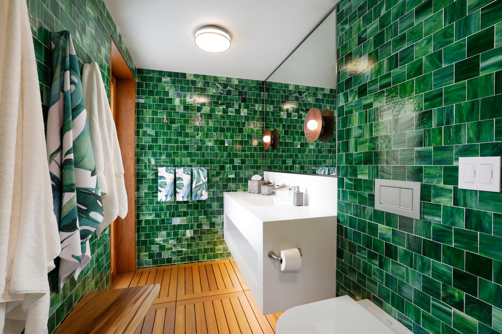 Photo of a mid-sized beach style 3/4 bathroom in Orange County with white cabinets, green tile, medium hardwood floors, an integrated sink, brown floor, white benchtops, open cabinets, a wall-mount toilet, green walls and solid surface benchtops.