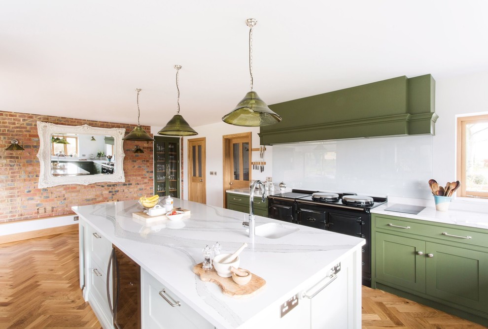 This is an example of a large country u-shaped open plan kitchen in Sussex with a farmhouse sink, beaded inset cabinets, green cabinets, quartzite benchtops, glass sheet splashback, black appliances, medium hardwood floors, with island, brown floor and white benchtop.